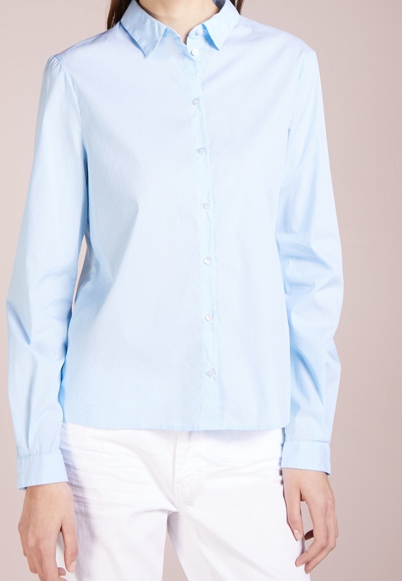 Chemise bleu clair à manches longues avec boutons, portée avec un pantalon blanc, visible du cou aux hanches sur un fond uni.