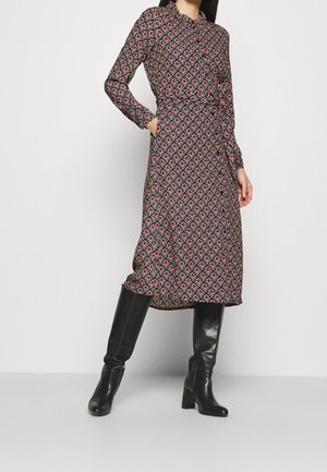 Schwarzes Kleid mit langen Ärmeln, rotem und weißem Blumenmuster, tailliert und mit Taschen. Kombiniert mit kniehohen schwarzen Stiefeln für einen Kontrast.