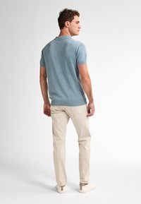 Světlomodré polo tričko s krátkým rukávem a hladkou texturou. V kombinaci se světlobéžovými kalhotami a bílými botami, otočen mírně od kamery.