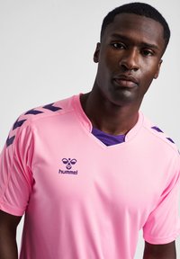 Rosa Sporttrikot mit lila Kragen, das schwarze Chevron-Akzente an den Schultern und ein Logo in lila Schrift zeigt. Leichtes Material.