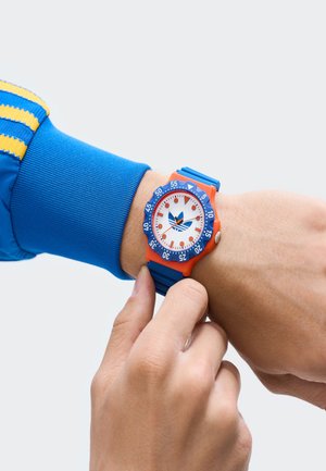 Barvit ura s pomarančno ohišjem, modrim bezelom in belim ciferblatom. Ima moder gumijast pas in adidasovo logotip na sredini ciferblata.