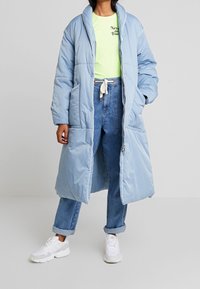 Cappotto lungo imbottito azzurro chiaro con bottoni a pressione, ampio colletto e due tasche laterali; indossato su una t-shirt verde e jeans blu. Sneakers bianche.