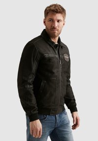 Man draagt een zwart jack met rits en een patch op de borst en blauwe spijkerbroek, kijkt naar links tegen een effen achtergrond.