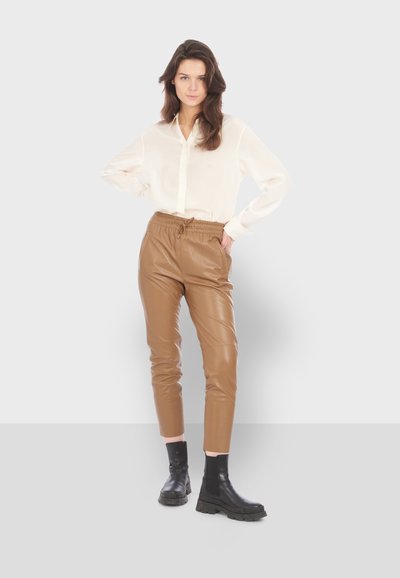 Chemise à col crème, pantalon en cuir élastique marron à taille haute et bottes noires à semelle épaisse. La tenue présente un design élégant et ajusté.