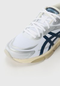 Pantofii sport albi cu accente argintii, textură din plasă, detalii ale logo-ului în nuanță de navy și talpă groasă de culoare crem. Șireturile sunt drepte și albe.