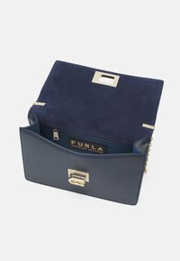 Borsa a mano in pelle blu con patta in camoscio, chiusura in tono oro e tracolla a catena. L'interno include una tasca con zip e un'etichetta con logo.