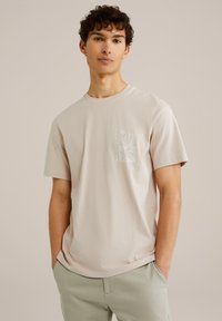 Camiseta de manga corta color beige claro, hecha de algodón suave, que presenta un diseño gráfico en el lado izquierdo del pecho con la palabra "PARADISE" y motivos de plantas.