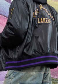 Chaqueta bomber de satén negro con bordado de "Los Angeles Lakers" en púrpura y amarillo. Cuenta con un dobladillo acanalado con rayas moradas.