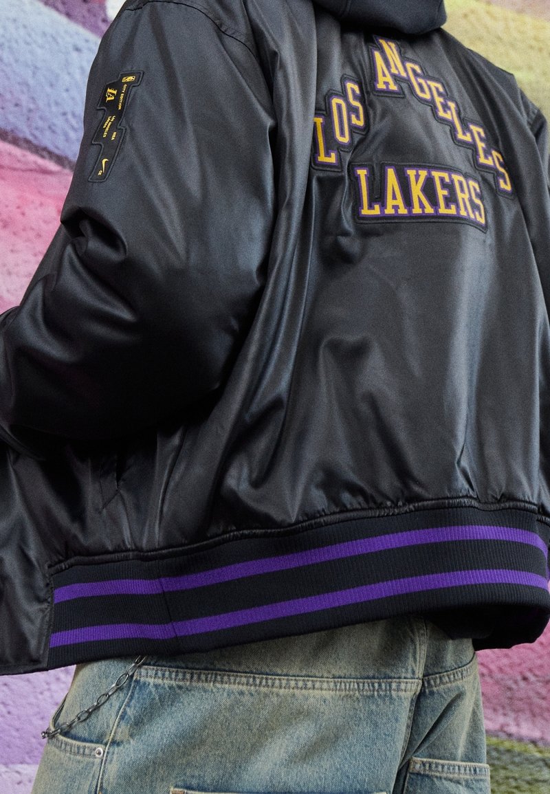 Chaqueta bomber de satén negro con bordado de "Los Angeles Lakers" en púrpura y amarillo. Cuenta con un dobladillo acanalado con rayas moradas.