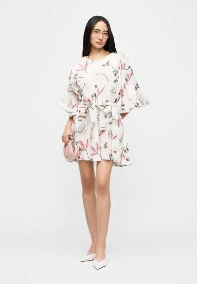 Φλοράλ φόρεμα με κουκούλα και μανίκια κουδούνια, σε κρεμ και ροζ αποχρώσεις. Διαθέτει ζώνη στη μέση και βελούδινη υφή. Το μοντέλο κρατάει μια μικρή ροζ τσάντα.