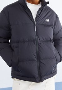 Mężczyzna w czarnej pikowanej kurtce New Balance zapiętej na biały długi rękaw, z rękami w kieszeniach kurtki.