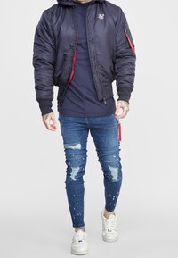 Chaqueta bomber negra con forro rojo, detalles de cremallera y bolsillos laterales, combinada con jeans azules desgastados y zapatillas blancas.