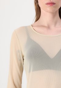 Gjennomsiktig langermet topp i beige mesh stoff, med scoop-hals og subtile mønsterdetaljer som ligner et trekantdesign under.
