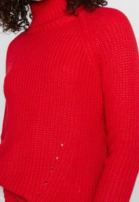 Pull rouge en maille avec un col roulé haut. Présente un motif texturé et de petits trous, offrant une coupe décontractée et des poignets côtelés.