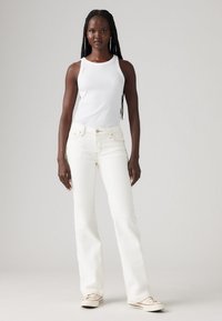 Hvit tanktopp kombinert med hvite utsvingte jeans, med kontraststing og avslappet passform. Skoene er i kanvas med svake mønsterdetaljer.