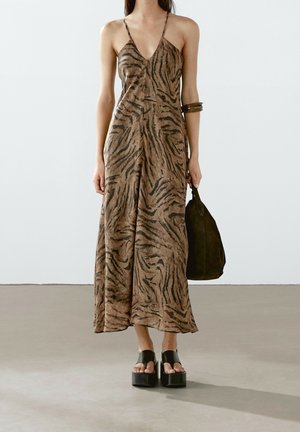 Kobieta w brązowej sukience na ramiączkach w tygrysie paski, czarnych sandałach na platformie, trzymająca ciemnozieloną torebkę z zamszu i nosząca brązową bransoletkę.