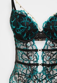 Črni čipkasti modrček z turkizno cvetlično vezenino, strukturirane oblike, nastavljivimi naramnicami in zlato kovinskimi poudarki. Polprosojen dizajn z večplastnimi teksturami.