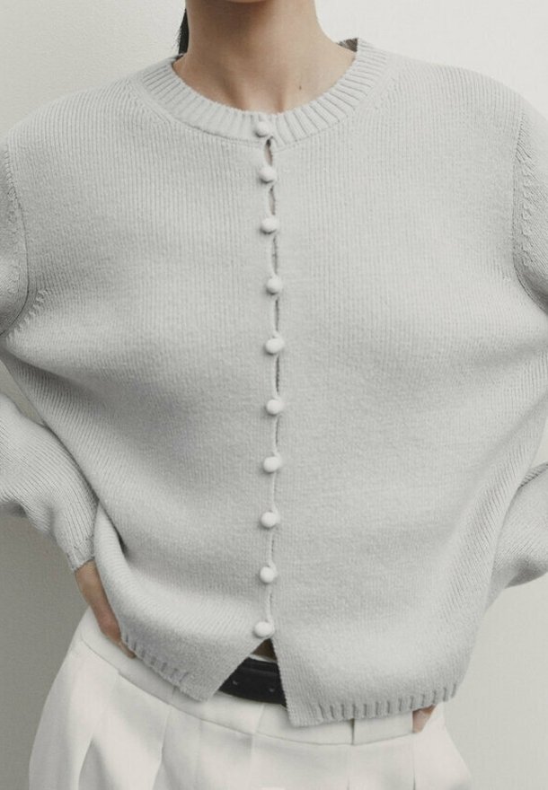 Femme portant un cardigan en maille gris clair avec des boutons en pompon texturés centrés, associé à un pantalon blanc taille haute.