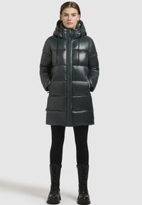 Polstret mørkegrøn parka, glat materiale, knælængde, med hætte og snore, lynlås-lommer, parret med sorte leggings og kampstøvler.