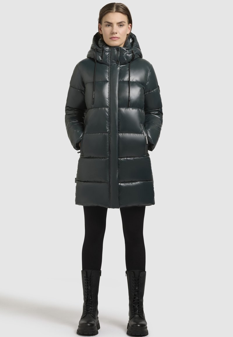 Polstret mørkegrøn parka, glat materiale, knælængde, med hætte og snore, lynlås-lommer, parret med sorte leggings og kampstøvler.