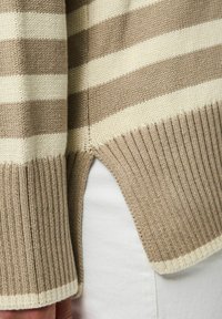 Beżowy sweter w paski w kolorze kremowym z prążkowanymi mankietami i dołem. Posiada boczne rozcięcia dla dodatkowego stylu. Widoczna tekstura dzianiny.