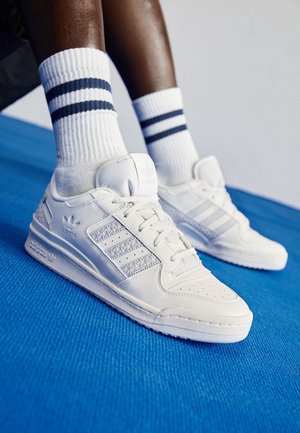 Benendelen met witte Adidas-sneakers en witte sokken met twee zwarte strepen, staand op een felblauwe textuurrijke ondergrond.