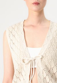 Knit cardigan in light beige with a diamond pattern, open front, and a tied closure. Lightweight texture. Sleeveless design.