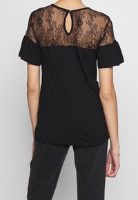 Kvinna klädd i svart kortärmad topp med genomskinlig blommig spets vid axlarna och knappöppning på övre ryggen, kombinerad med mörka byxor.