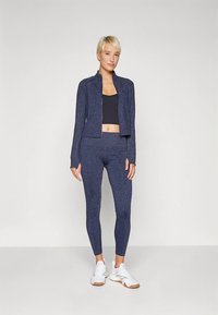 Sportovní souprava v námořnické modři zahrnuje přiléhavou bundu na zip a legíny s vysokým pasem, obojí z texturované látky, doplněné bílými teniskami.