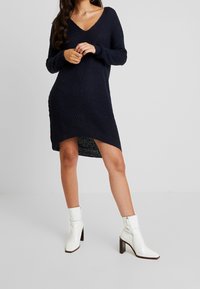 Marinblå stickad klänning med djup V-ringning, ribbat mönster och sidoklyfta, kombinerad med vita ankelboots med klackar.