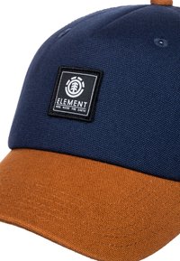 Cappello da baseball blu navy e arancione bruciato con una patch nera e bianca del logo Element che presenta un albero e la scritta: "Vento Acqua Fuoco Terra."
