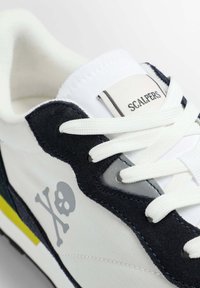 Sneakers bianchi con accenti in suede navy, lacci bianchi e logo a forma di teschio grigio. Presentano un colletto imbottito e una suola testurizzata. Etichetta del brand visibile.