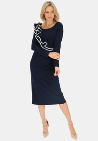 Granatowa sukienka z długimi rękawami i dopasowanym krojem. Posiada biały falbaniasty akcent na jednym ramieniu. Gładka tekstura materiału, długość do kolan.