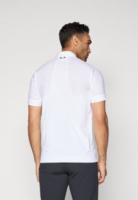 Biała koszulka polo z krótkim rękawem i kołnierzykiem, z tyłu detal z logo oraz gładkim, lekkim materiałem. Prosty, elegancki design.