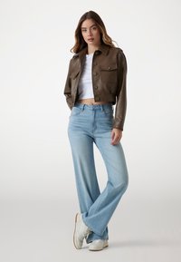 Giubbotto di pelle marrone a maniche corte con due tasche frontali, abbinato a jeans a campana azzurri e sneakers bianche. Texture liscia e design aderente.