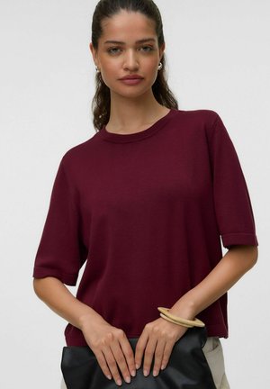 Kvinde iført en burgunder kortærmet top, der holder en sort tekstureret clutch-taske, med et lyst træarmbånd på håndleddet.