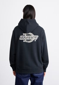 Sort hættetrøje med et stort hvidt "Dickies" logo på ryggen. Har snoretræk i hætten, ribbet manchetter og en blød stofoverflade.