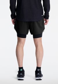 Hombre con pantalones cortos de running negros sobre mallas de compresión negras, calcetines negros de New Balance y zapatillas de running negras, de pie sobre un fondo blanco.