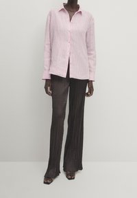 Personne portant une chemise à boutons rayée rose et blanche, un pantalon large noir texturé et des sandales noires à doigts de pied ouverts, sur un fond uni.