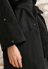Svart regnrock med texturerad yta, vida ärmar och knytbälte. Har knappar på ärmsluten och en hand som bär en ring.