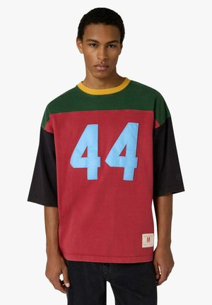 Giovane uomo che indossa una maglietta a blocchi di colore con sezioni verdi, rosse e nere, grande numero blu "44" sul davanti e bordo giallo al collo.