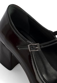 Chaussure Mary Jane en cuir verni noir avec un talon block et une sangle en boucle argentée, présentée sous un angle rapproché.