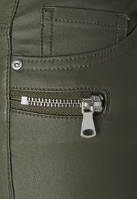 Olijfgroen stof met een gladde textuur heeft een klein ritszakje, zilveren rits hardware en contrasterend stiksel langs de randen.