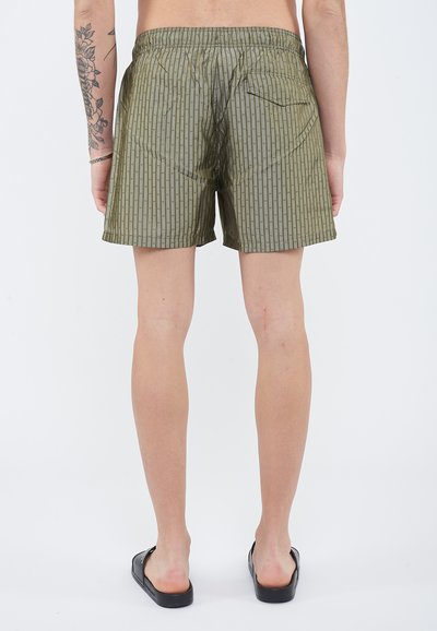 Άνδρας που στέκεται ξυπόλητος φορώντας πράσινα σορτς με σχέδιο και μαύρα σανδάλια, εμφανές τατουάζ στο αριστερό χέρι, θέα από πίσω.