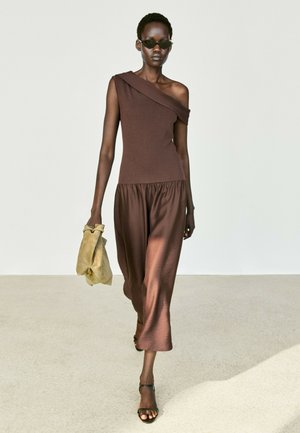 Frau trägt braunes Ein-Schulter-Kleid, dunkle Sonnenbrille, schwarze Sandalen und hält eine beige Handtasche und geht auf einer hellen Oberfläche.