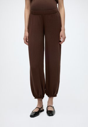 Kobieta nosząca brązowe spodnie o luźnym kroju z elastycznymi mankietami, w połączeniu z czarnymi płaskimi butami na jasnym, jednolitym tle.