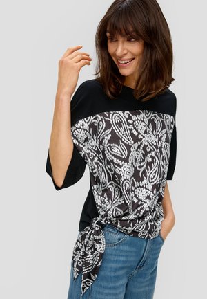 Haut noir à manches courtes avec un motif paisley blanc. Il présente un détail noué à la taille et une coupe ample. Associé à un jean bleu.