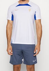 Camiseta deportiva blanca de Nike con acentos azules, que presenta un logo dorado y un emblema francés. Combinada con pantalones cortos atléticos grises, de tela suave.