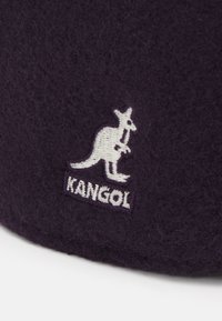 Cappello di lana viola con una consistenza morbida, caratterizzato da un logo di un canguro ricamato in bianco e dal nome del marchio "KANGOL" sottostante.