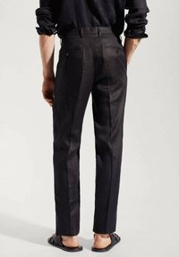 Person wearing tailored black pants, hands relaxed at sides, paired with rolled-up black shirt sleeves and black woven sandals.